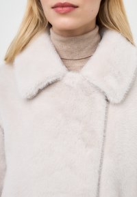 Cappotto in pelliccia sintetica di un leggero beige con un grande colletto, caratterizzato da una morbida texture e una lieve lucentezza. Indossato sopra un dolcevita a coste beige.