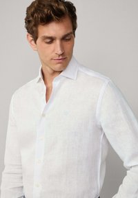 Jonge man kijkt omlaag en draagt een wit linnen overhemd met lange mouwen en een kleine geborduurde logo op de borst.