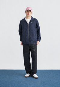 Νεαρός άνδρας που στέκεται φορώντας ναυτικό μπλε μπουφάν με φερμουάρ και σχέδια, λευκό μπλουζάκι, μαύρο τζιν με τρουκς, λευκά αθλητικά παπούτσια και ροζ καπέλο σε μπλε χαλί.