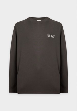 Homme portant un t-shirt noir à manches longues avec texte, pantalon clair, casquette beige et baskets beiges, debout avec une main dans la poche.
