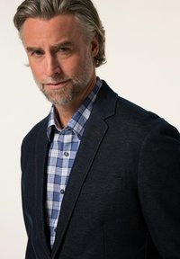 Marinblå strukturerad kavaj över en blå och vit rutig skjorta. Kavajen har en slagkrage och en enkel bröstficka med mörka knappar.