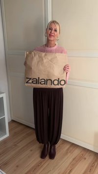 Bolsa de papel marrón para comprar con "zalando" impreso en letras negras en negrita; combinada con una blusa rosa claro y pantalones oscuros de corte holgado.
