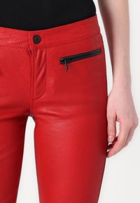 Red perforated leather pants feature a sleek design with a side zip pocket and a smooth finish, accentuated by a black zipper detail.