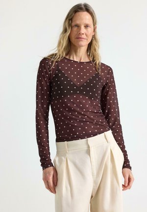 Keski-ikäinen nainen, joka pukee päällensä läpikuultavan ruskean, pilkullisen pitkähihaisen paidan mustan alusvaatteen kanssa ja korkea vyötäröiset kermaiset, laskostetut housut, seisomassa.