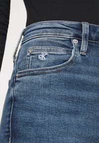 Primer plano de un bolsillo de jeans de mezclilla azul con el logotipo "cK" cosido, remaches de metal y un lazo para el cinturón, combinado con una camiseta negra.