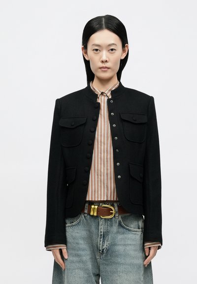 Sort skræddersyet jakke med høj krave, med to flade brystlommer og knaplukning. Bæres over en stribet skjorte og slidte jeans.