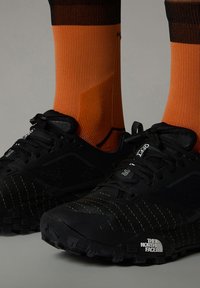 Svarte atletiske sko med teksturerte mesh-paneler og gule sømdetaljer, kombinert med lyse oransje sokker med en svart stripe øverst.