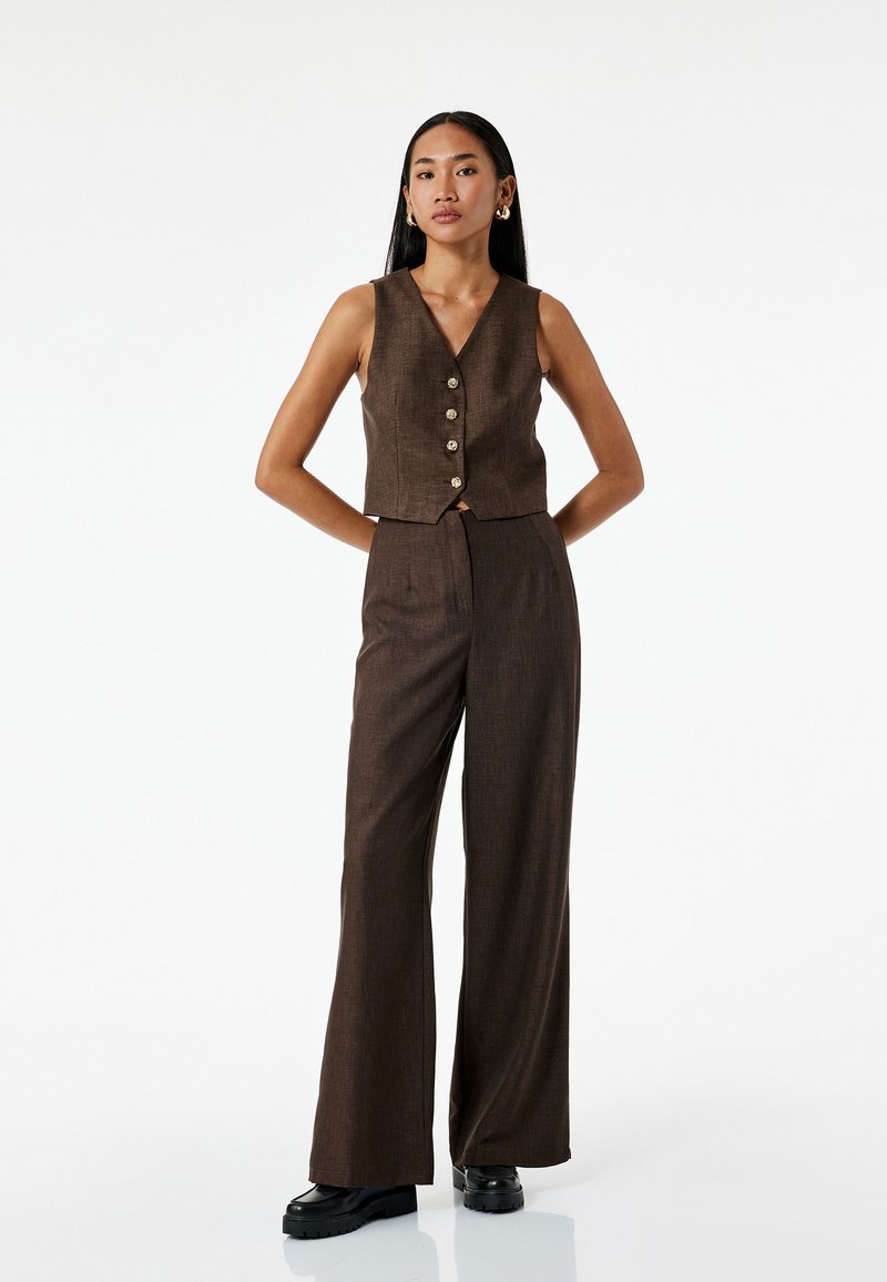 Donkerbruine cropped vest met knoopdetails, gecombineerd met wijde broek van een textiel met structuur en een getailleerde pasvorm. Zwarte platform schoenen.