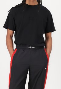 Osoba oblečená v černém tričku s krátkým rukávem a černých teplácích Adidas s červenými bočními pruhy a bílými logy, s rukama v kapsách.