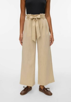 Pantalons larges beiges en tissu texturé, avec une taille à nouer et des plis avant. Portés avec un débardeur noir et des chaussures marron.