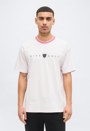 Jaunas vyras, dėvintis baltą Nike Golf marškinėlį su rožiniu apykakle, stovintis prieš šviesią vientisą fono sieną, žiūrintis į priekį.