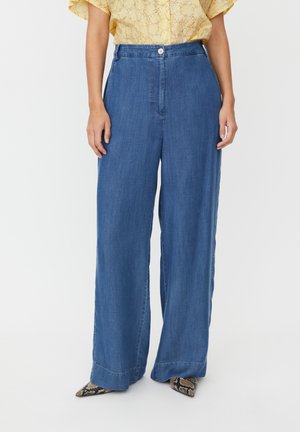 Pantalon en jean à jambes larges de teinte bleu moyen, avec une fermeture à boutons sur le devant et une texture lisse, assorti à un haut jaune imprimé et des chaussures à motifs.