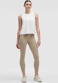 Fehér, ujjatlan, laza szabású felső, bézs, magas derekú leggings-szel kombinálva. A modell semleges háttér előtt áll, világos sportcipőt visel.