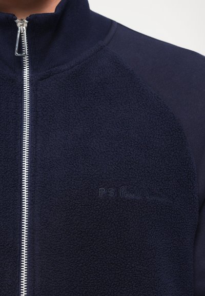 Jachetă navy cu fermoar, confecționată din material de fleece, cu guler înalt și un panou lateral neted, având un logo discret brodat pe piept.