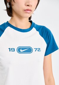Lühikeste varrukatega T-särk, mille keha on valge ja raglanvarrukad sinised. Ees on sinine Nike logo ja "1972" graafika.