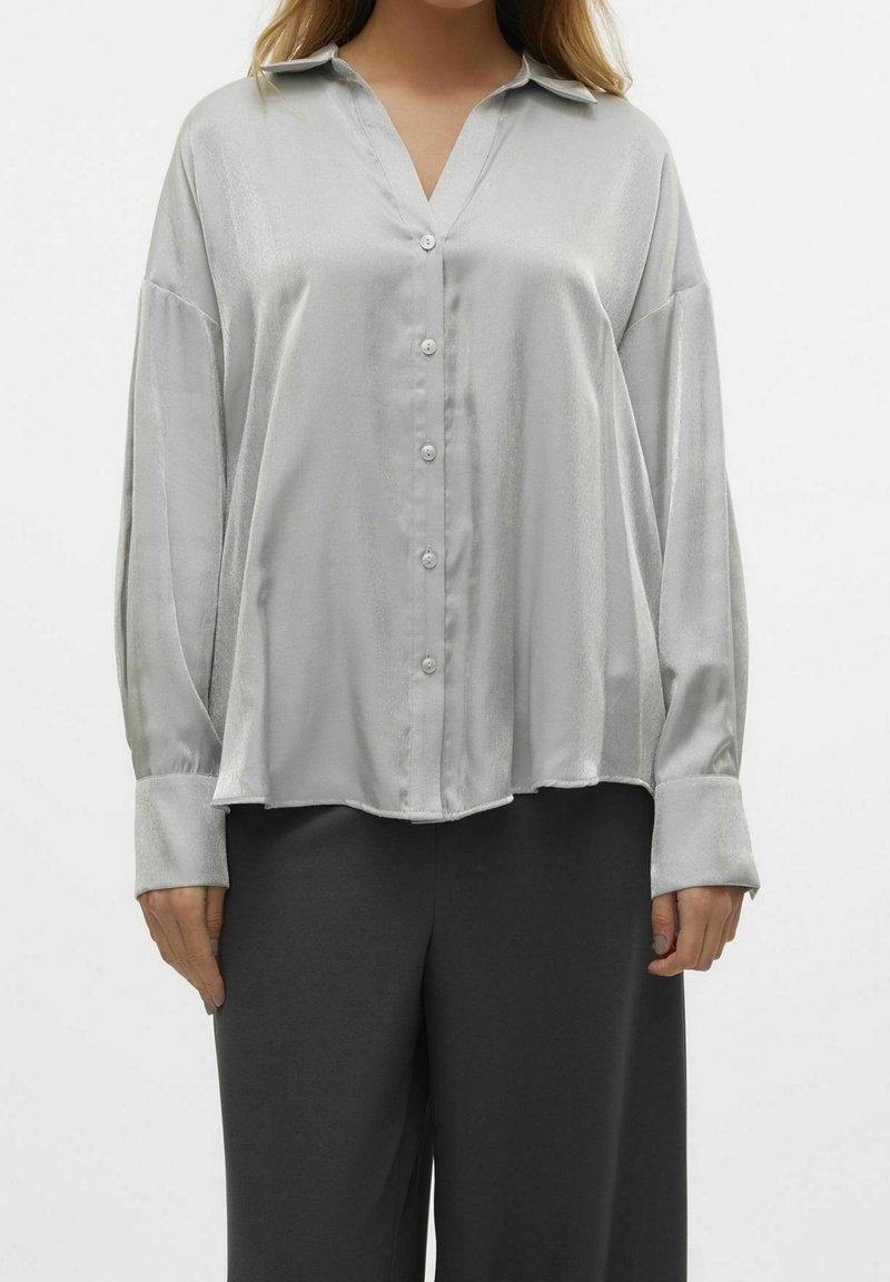Femme portant un chemisier ample en satin gris clair à boutons, manches longues et larges poignets, associé à un pantalon noir.