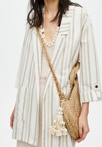 Striped, beige linen blazer with two pockets. Worn over a knitted top, paired with a woven straw bag featuring decorative tassels.
