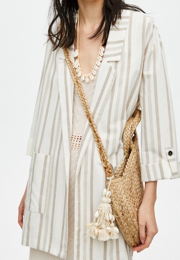 Striped, beige linen blazer with two pockets. Worn over a knitted top, paired with a woven straw bag featuring decorative tassels.