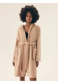 Beżowy szlafrok z polaru z kapturem, długimi rękawami, dwoma przednimi kieszeniami i pasującym paskiem. Tekstura wydaje się miękka i przytulna.
