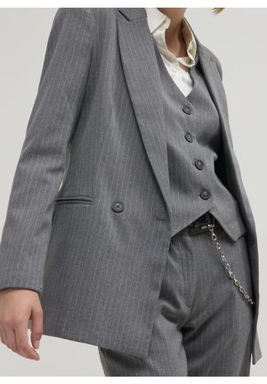 Person trägt einen grauen Nadelstreifanzug bestehend aus Blazer, geknöpfter Weste und Hose mit silberner Ketten-Accessoire und weißem Hemd mit Kragen.