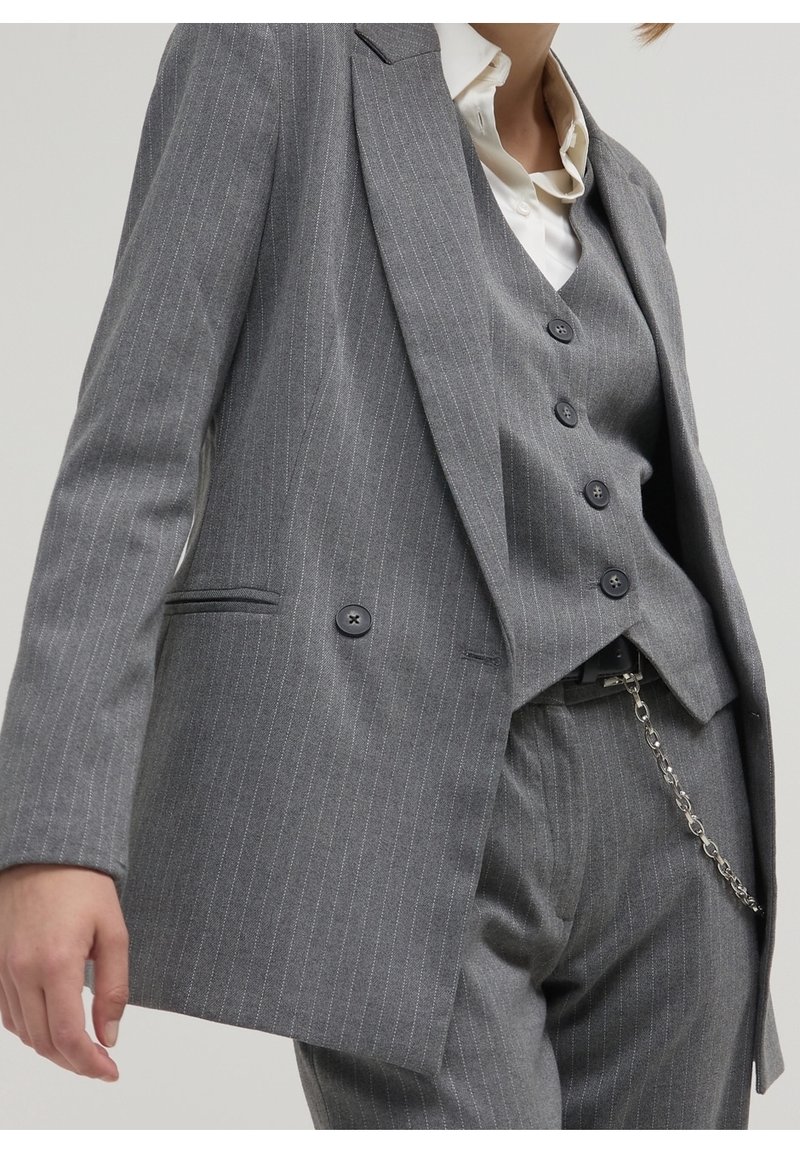 Personne portant un costume gris à rayures fines comprenant une veste, un gilet boutonné et un pantalon avec un accessoire chaîne argentée et une chemise blanche à col.