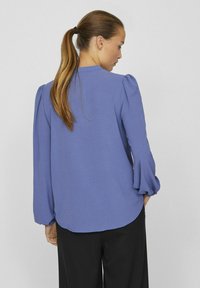 Blusa viola con maniche a sbuffo, scollo rotondo, tessuto testurizzato e orlo curvo. Indossata con pantaloni neri.