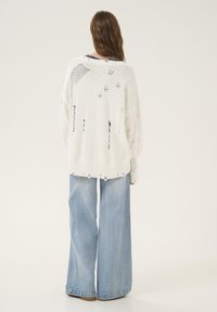 Witte gebreide oversized trui met een versleten uitstraling, gecombineerd met lichtblauwe wijde jeans. Zachte textuur met geribbelde boorden en zoom.