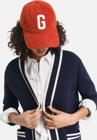 Red corduroy cap with a white letter "G" at the front, worn with a navy cardigan featuring white trim, button detailing, and a collared shirt.