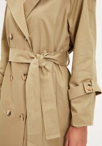 Béžový trenčkot s dvouřadým zapínáním na knoflíky, páskem v pase a manžetami na knoflíky na rukávech. Hladká tkanina s klasickým designem.
