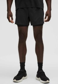 Pantaloni sportivi neri realizzati in un materiale leggero, con una texture liscia e dettagli in rete laterali, abbinati a sneakers nere e calze.