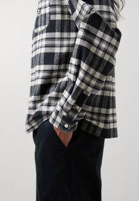 Personne portant une chemise à carreaux noir et blanc à manches longues avec boutons, main dans la poche, et pantalon noir, debout devant un fond uni.