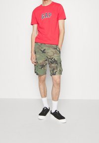 Persona que lleva una camiseta roja de GAP, pantalones cortos de camuflaje, calcetines blancos y zapatillas negras, de pie con una mano en el bolsillo.