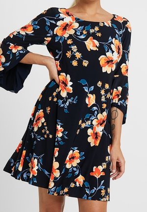 Czarna sukienka z pomarańczowo-kremowym kwiatowym wzorem, rozkloszowanymi rękawami i spódnicą w kształcie litery A, noszona przez osobę z tatuażem na przedramieniu.