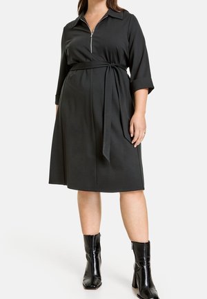 Woman wearing knee-length black dress with three-quarter sleeves, a tie belt, front zipper, and black heeled ankle boots.