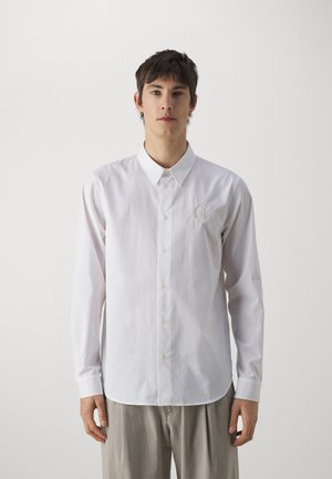 Camicia bianca a maniche lunghe con bottone frontale, colletto a punta e ricamo logo discreto sul lato sinistro del petto; tessuto morbido e vestibilità su misura.