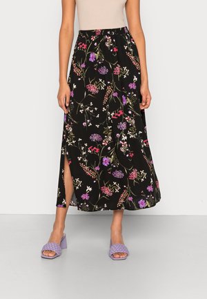 Jupe longue noire à fleurs avec fente latérale portée avec un haut beige et des sandales à talon carré lavande sur un fond clair uni.