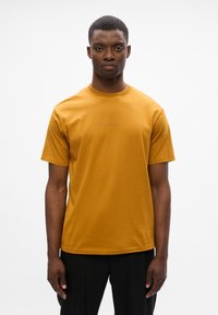 Lyhythihainen sinapinkeltainen t-paita puuvillasekoitetta, jossa on pyöreä kaula-aukko ja hienovarainen brändäys edessä. Sileä pinta, rento malli.