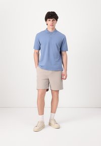Lyseblå pikéskjorte med krage, korte ermer og to knapper. Beige stripete shorts. Hvite sokker og beige sko. Enkelt bakgrunn.