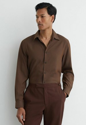 Uomo che indossa una camicia marrone a righe con bottoni infilata in pantaloni marrone scuro, in piedi con una mano in tasca su uno sfondo bianco.