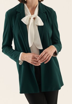 Kobieta ubrana w ciemnozieloną marynarkę na białą bluzkę z dużą zawiązaną kokardą przy szyi, stoi z rękami złożonymi z przodu.