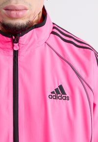 Pink Adidas zip-up jacket with black branding. Features black stripes along the shoulders and a high collar. Smooth, lightweight material.