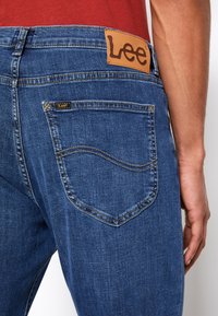 Blå denim jeans med et lærmerke i livet, akkompagnert av distinkt søm og en lomme med en buet design.