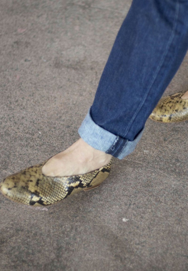 Chaussures avec imprimé serpent jaune et noir portées avec un jean bleu foncé retroussé sur un sol recouvert de moquette grise.