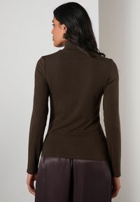 Brown long-sleeve top with a mock neck, ribbed texture, and fitted shape, paired with loose, shiny dark pants.