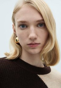 Bež in rjav pulover z teksturiranim pletenim vzorcem. Model ima ravne blond lase in nosi velike zlate uhane. Nevtralna ozadje.