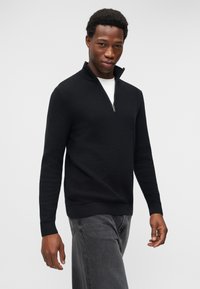 Άνδρας που φοράει μαύρο υφασμάτινο πουλόβερ με φερμουάρ έως τη μέση πάνω από λευκό πουκάμισο και ξεβάμμένα μαύρα τζιν, στέκεται μπροστά σε απλό ανοιχτό φόντο.