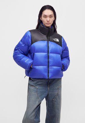 Човек, носещ синьо-черно подплатено яке North Face с ръце в джобовете и широки сини дънки на обикновен фон.