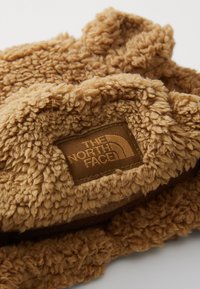Tecido fofo de cor caramelo com uma superfície texturizada. Apresenta um rótulo castanho com o logótipo "THE NORTH FACE". Design macio e peludo.