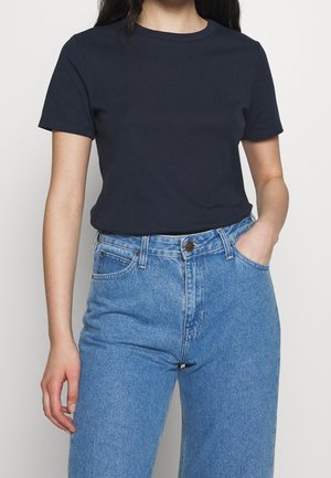 Frau trägt ein schlichtes schwarzes T-Shirt mit kurzen Ärmeln, in hoch taillierte blaue Jeans gesteckt, mit einer Hand in der Tasche.