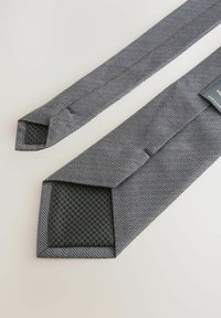 Gray herringbone tie with a textured black lining, featuring a pointed end and a small label sewn near the blade. Flat design, no embellishments.